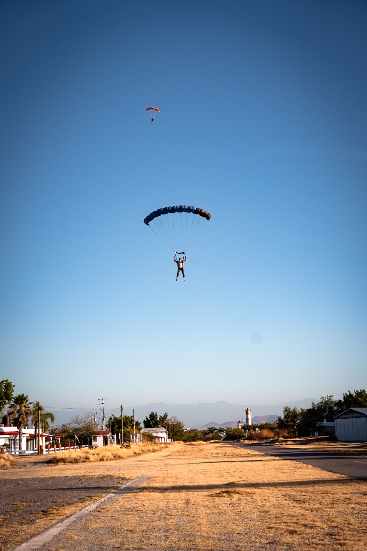 Skydiver flaring blue canopy for landing on a dirt strip with second jumper above