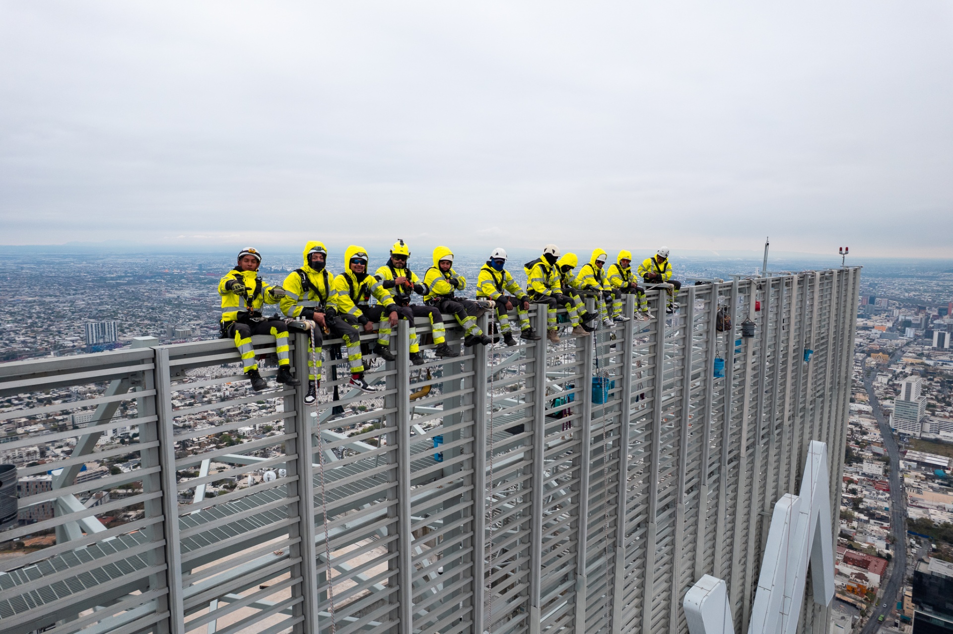 Aerial wide of high-altitude workers in hi-vis sitting on the steel facade of a Monterrey skyscraper