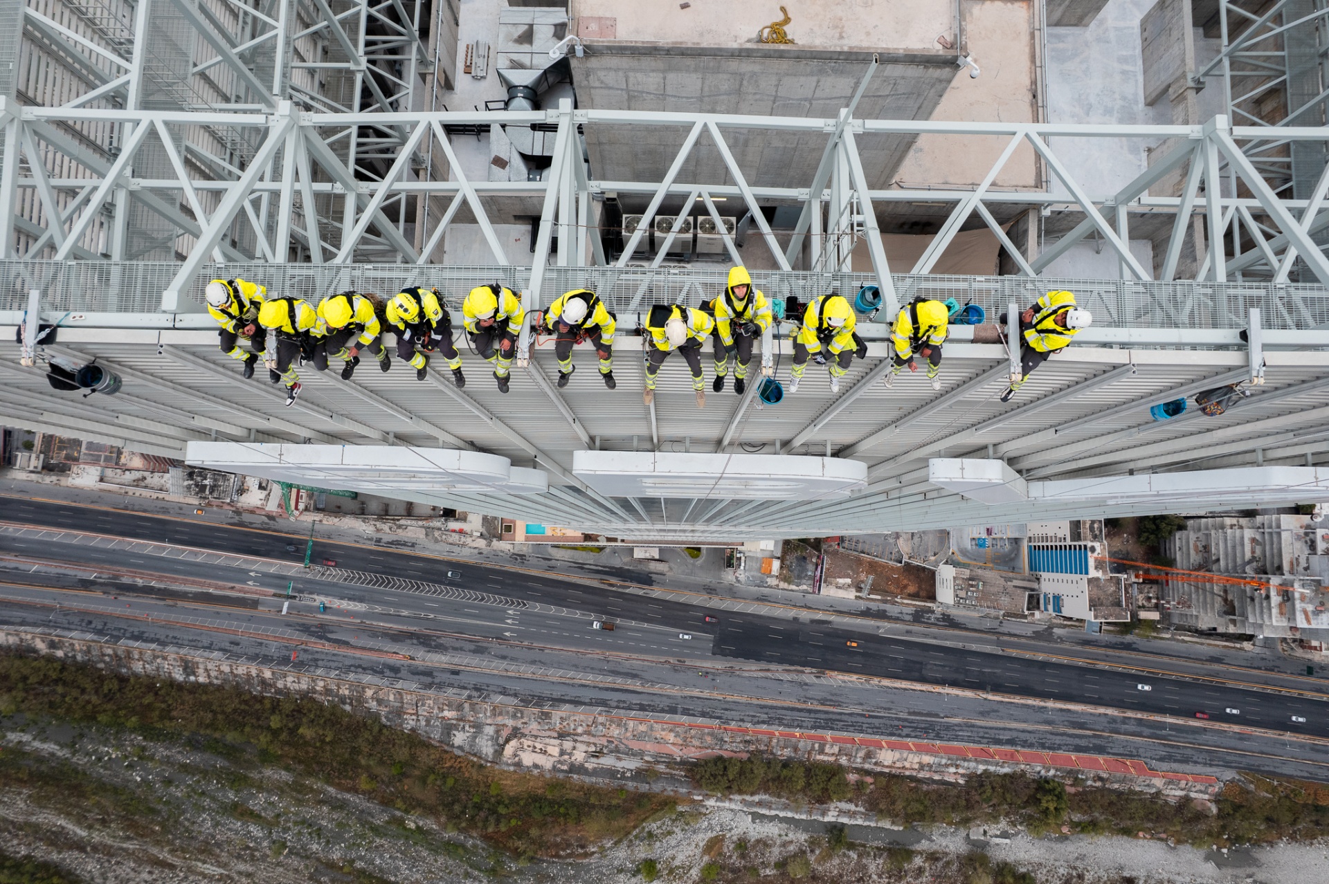 Top-down aerial of workers in yellow hi-vis sitting along a building's truss above the highway