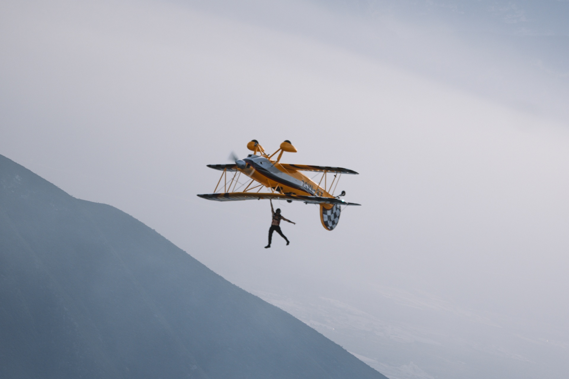 Wing walker hanging one-handed beneath biplane against mountainside