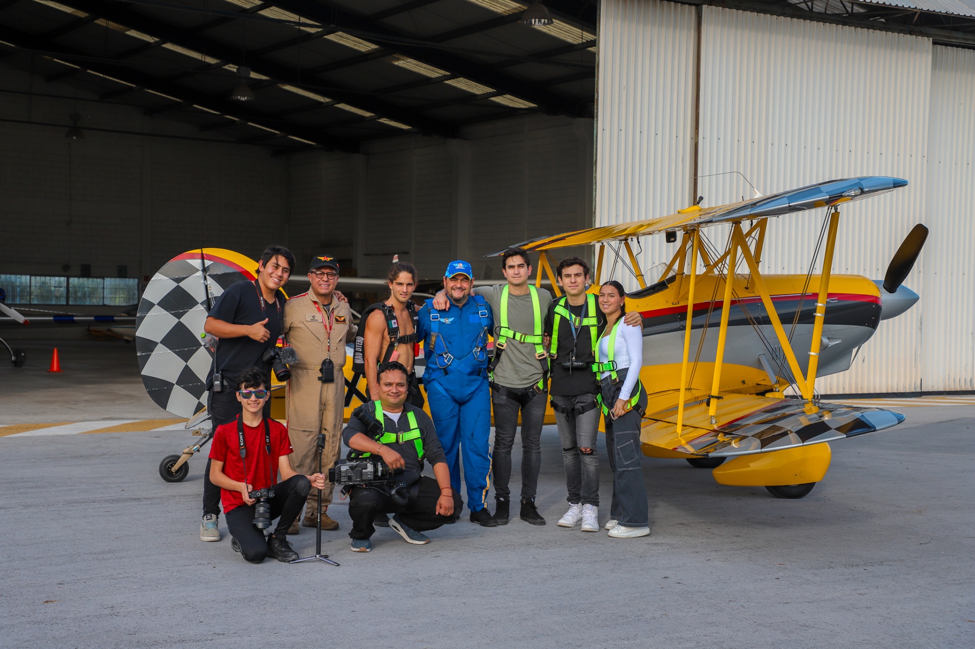 Full Dronebrothers crew posing with wing-walker and pilots in front of yellow Stearman biplane