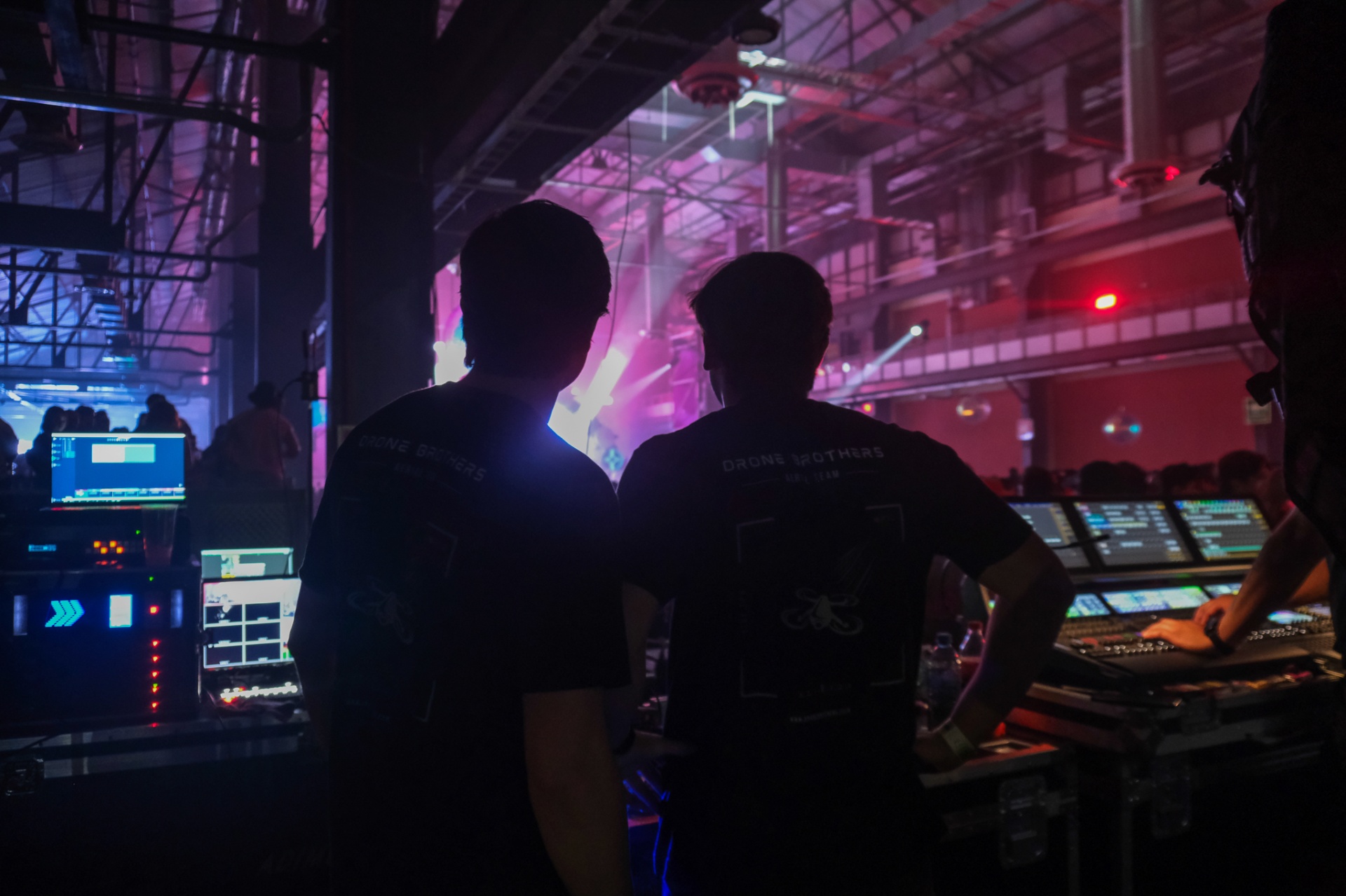 Two Drone Brothers operators silhouetted at FOH watching a stage washed in pink and purple light