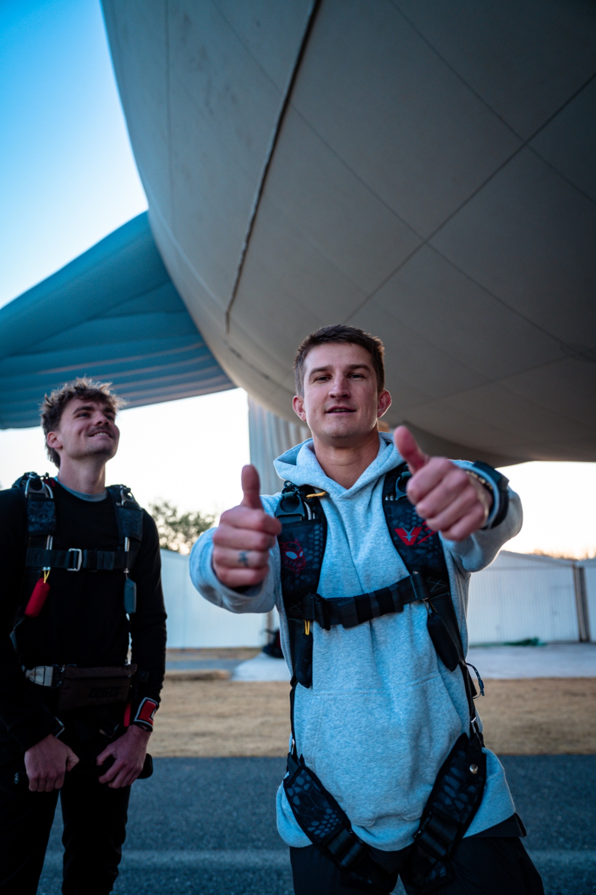 Skydiver in harness giving double thumbs up under a parked airship at dusk