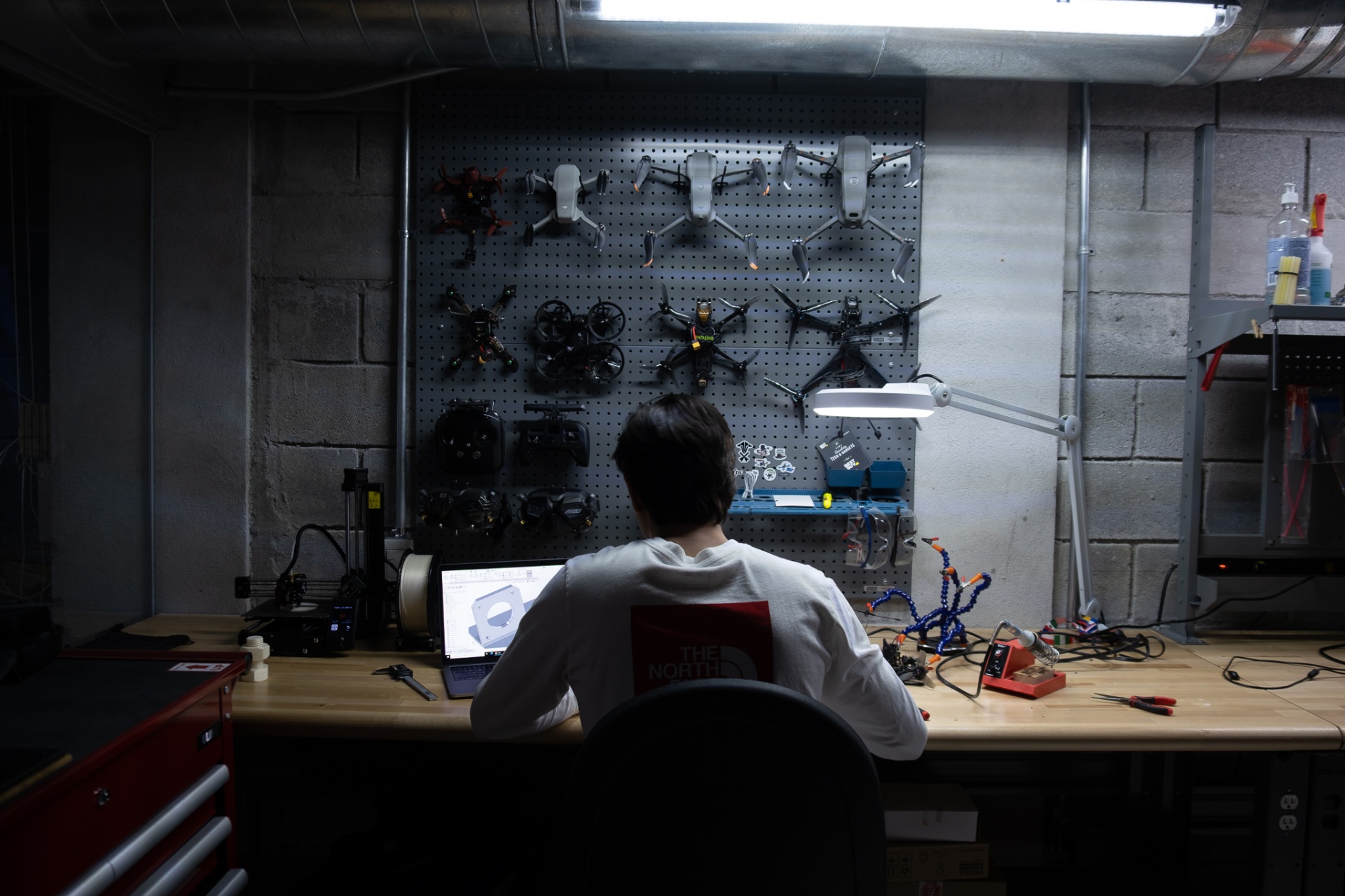 Operator at workshop bench in front of a pegboard wall lined with FPV quads, cinelifters, and DJI drones