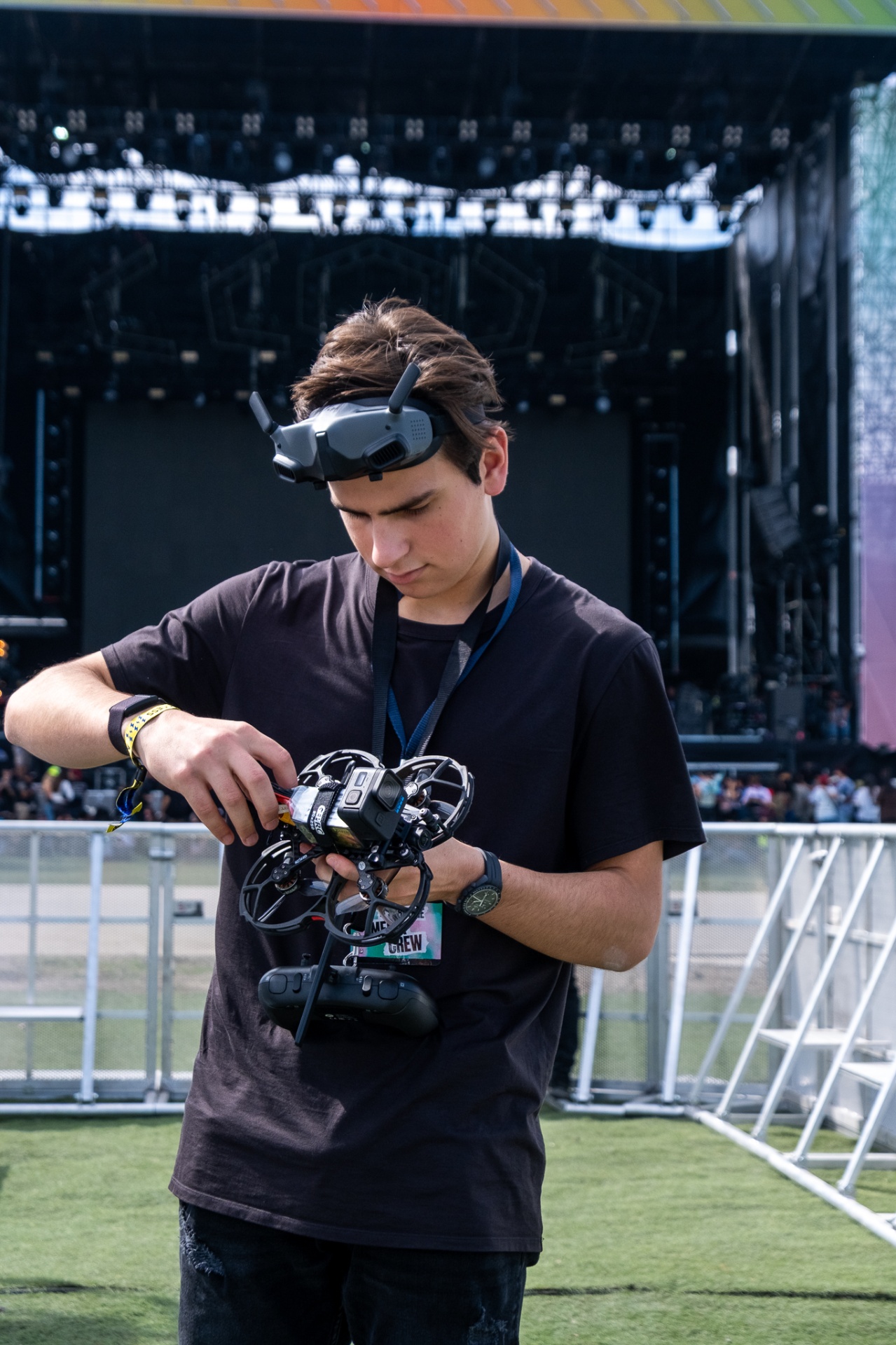 FPV pilot in crew tee plugging in a cinewhoop battery in front of a daylight festival stage