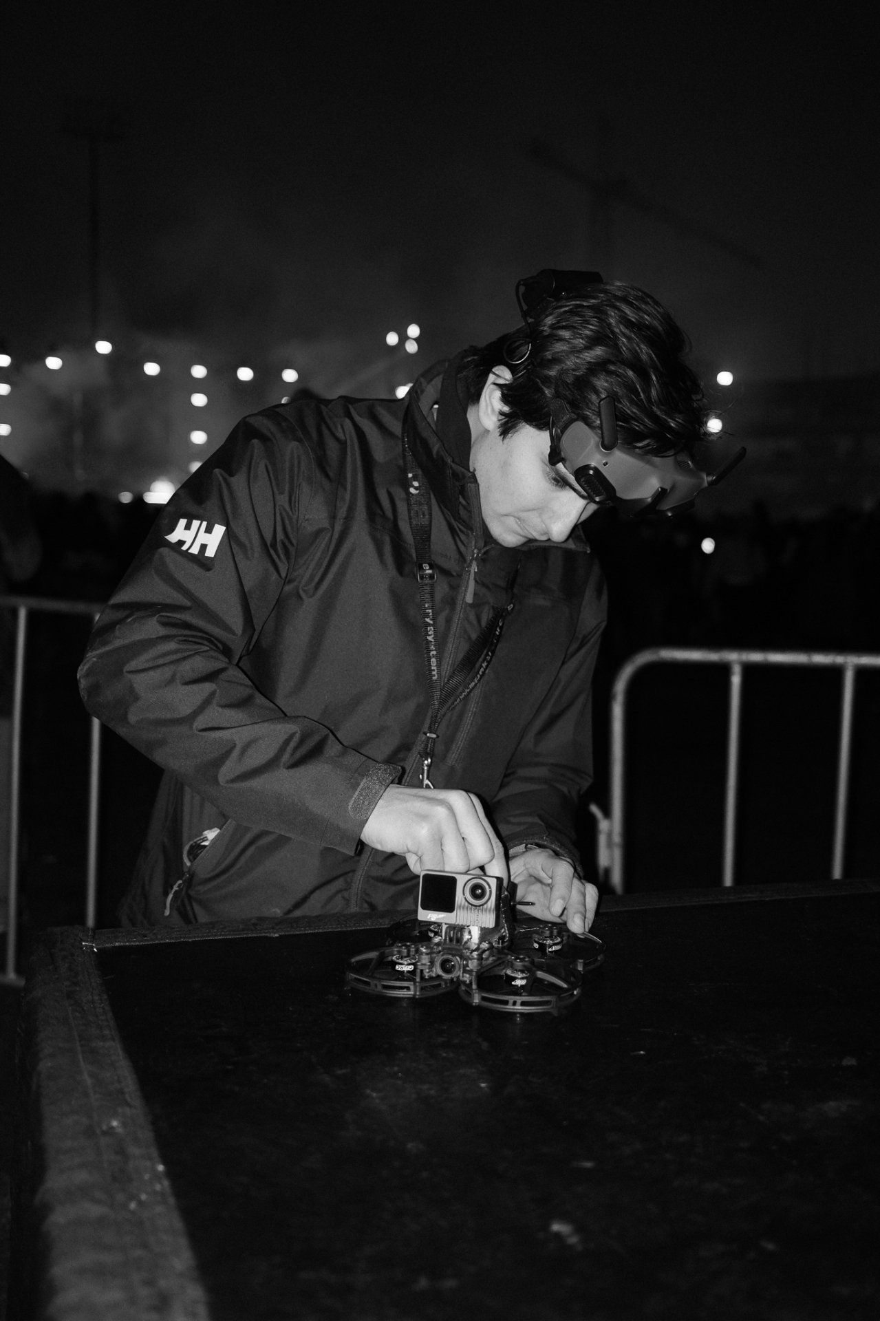 Black-and-white photo of FPV pilot prepping a cinewhoop with GoPro at a festival barricade at night