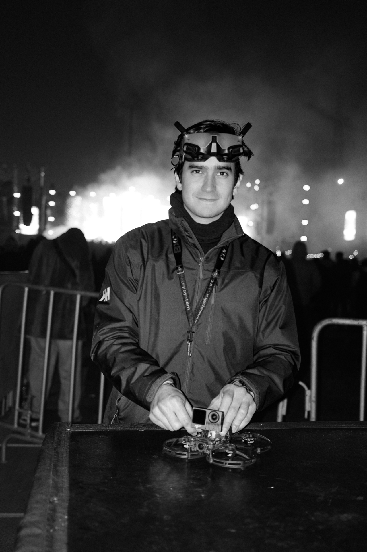Black-and-white portrait of FPV pilot with DJI goggles up, smoke and stage lights behind