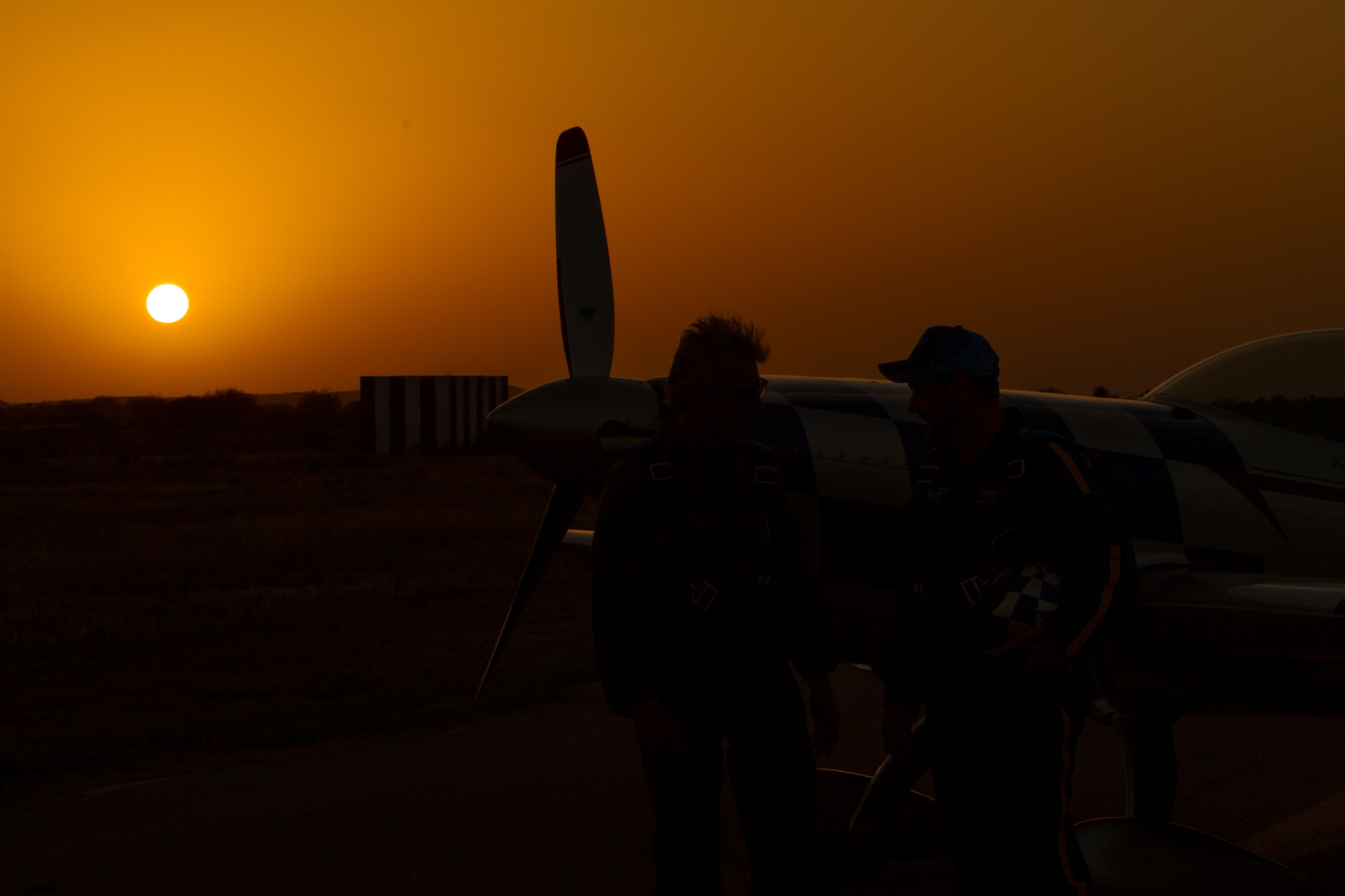 Silhouettes of two pilots beside an aerobatic plane with sun low on the horizon