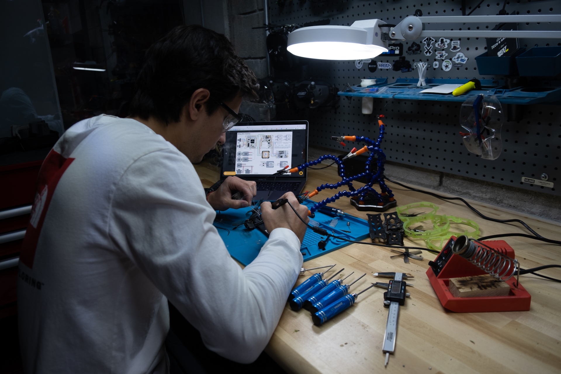 Builder in safety glasses soldering an FPV drone on a bench beside a wiring diagram and helping-hands